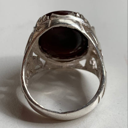 A silver filigree ring featuring a large, oval, dark reddish-brown gemstone, photographed on a plain light gray surface.