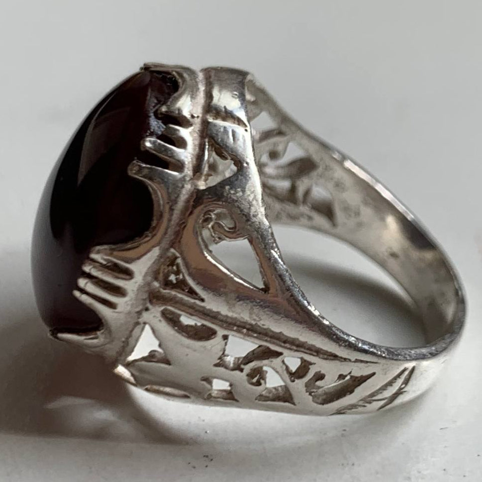 A silver ring with an ornate filigree band and a large, dark oval cabochon gemstone, photographed on a light gray surface.