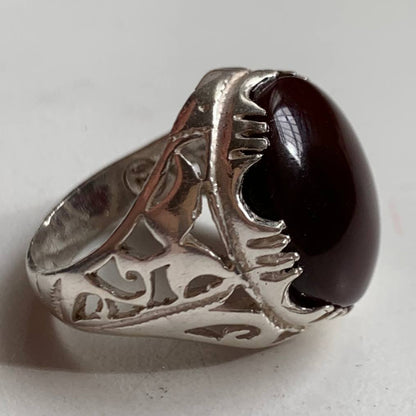 A silver ring with an intricate filigree band and a large, polished oval dark red gemstone, photographed on a light gray background.