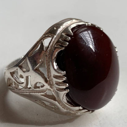 A silver ring featuring a large, polished oval dark red gemstone, photographed on a plain light gray surface.