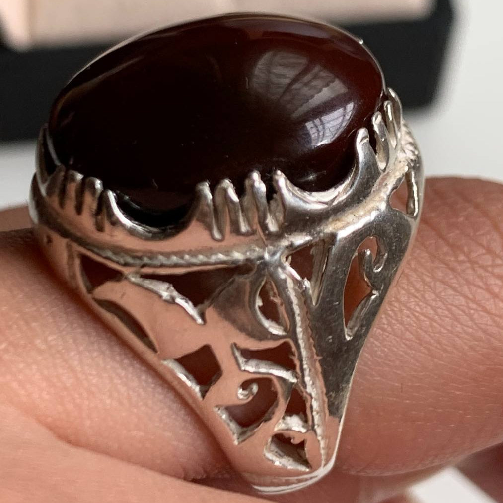 A silver ring featuring a large, polished oval cabochon gemstone in a deep reddish-brown color, displayed on a beige textured surface.