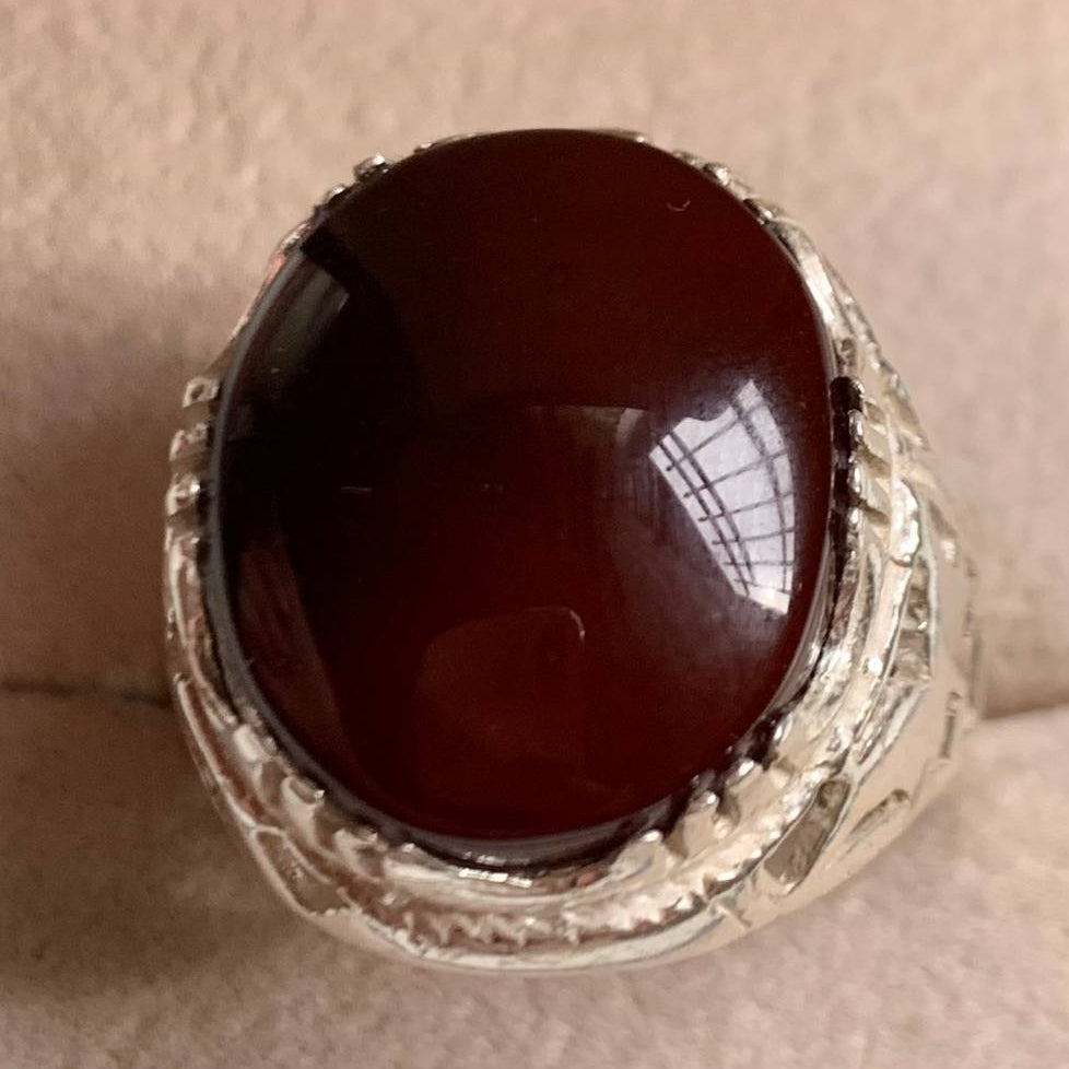 A silver ring featuring a large, oval, dark reddish-brown cabochon gemstone, photographed on a textured beige surface.
