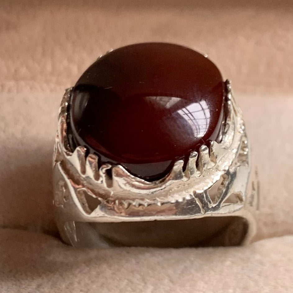 A silver-toned ring with a large, smooth, dark reddish-brown oval cabochon stone, displayed inside a beige velvet ring box.