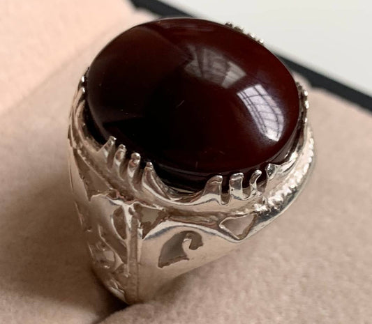 A silver ring with a large, polished dark reddish-brown cabochon gemstone, displayed on a beige textured ring box.